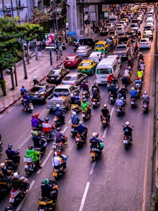 A congested urban street filled with numerous motorcycles and cars. Motorcycles dominate the foreground, each rider wearing a helmet and various colored clothing. Cars and taxis form a parallel line in the adjacent lane. Tall buildings and power lines frame the background, with a large concrete overpass stretching across the street.