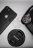 A flat lay of tech items including a black smartphone displaying the Apple logo, a PlayStation controller, an Apple logo in the center, and a Sony camera lens cap.