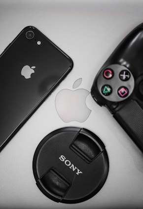 A flat lay of tech items including a black smartphone displaying the Apple logo, a PlayStation controller, an Apple logo in the center, and a Sony camera lens cap.