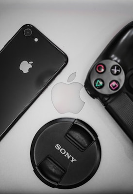 A flat lay of tech items including a black smartphone displaying the Apple logo, a PlayStation controller, an Apple logo in the center, and a Sony camera lens cap.