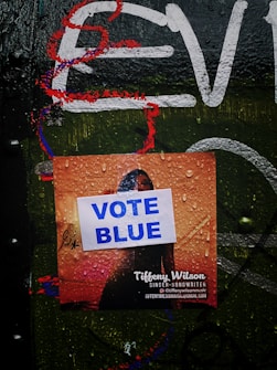 A damp poster is placed on a dark, graffiti-covered surface. The poster features text with the prominent phrase 'VOTE BLUE' printed on it. Beneath this, the name 'Tiffeny Wilson' is visible, identified as a singer-songwriter, along with social media and email contact information. Water droplets cover the entire surface, adding a wet appearance.