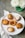 A variety of small, artisanal pies and tarts displayed on a white platter. Each pastry features different toppings and crust designs, showcasing intricate baking skills. The setting includes a green vase with white flowers and a striped cloth napkin on a gray tabletop.
