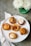 A variety of small, artisanal pies and tarts displayed on a white platter. Each pastry features different toppings and crust designs, showcasing intricate baking skills. The setting includes a green vase with white flowers and a striped cloth napkin on a gray tabletop.