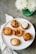 A variety of small, artisanal pies and tarts displayed on a white platter. Each pastry features different toppings and crust designs, showcasing intricate baking skills. The setting includes a green vase with white flowers and a striped cloth napkin on a gray tabletop.