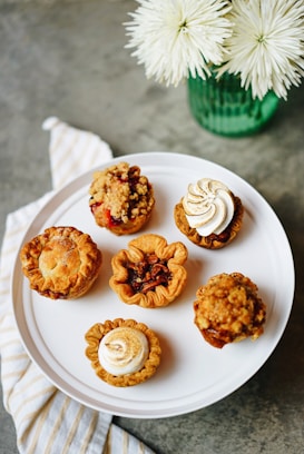 A variety of small, artisanal pies and tarts displayed on a white platter. Each pastry features different toppings and crust designs, showcasing intricate baking skills. The setting includes a green vase with white flowers and a striped cloth napkin on a gray tabletop.