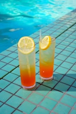 Two tall glasses filled with a colorful, layered beverage are placed on a tiled surface. Each drink is garnished with a slice of lemon on the rim. The background includes the edge of a swimming pool with clear blue water.