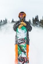 A person in winter gear holds a snowboard vertically in a snowy outdoor setting. They are wearing a black jacket, bright orange pants, yellow gloves, and a helmet with goggles. The background features snow-covered trees, creating a wintry atmosphere.