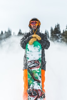 A person in winter gear holds a snowboard vertically in a snowy outdoor setting. They are wearing a black jacket, bright orange pants, yellow gloves, and a helmet with goggles. The background features snow-covered trees, creating a wintry atmosphere.
