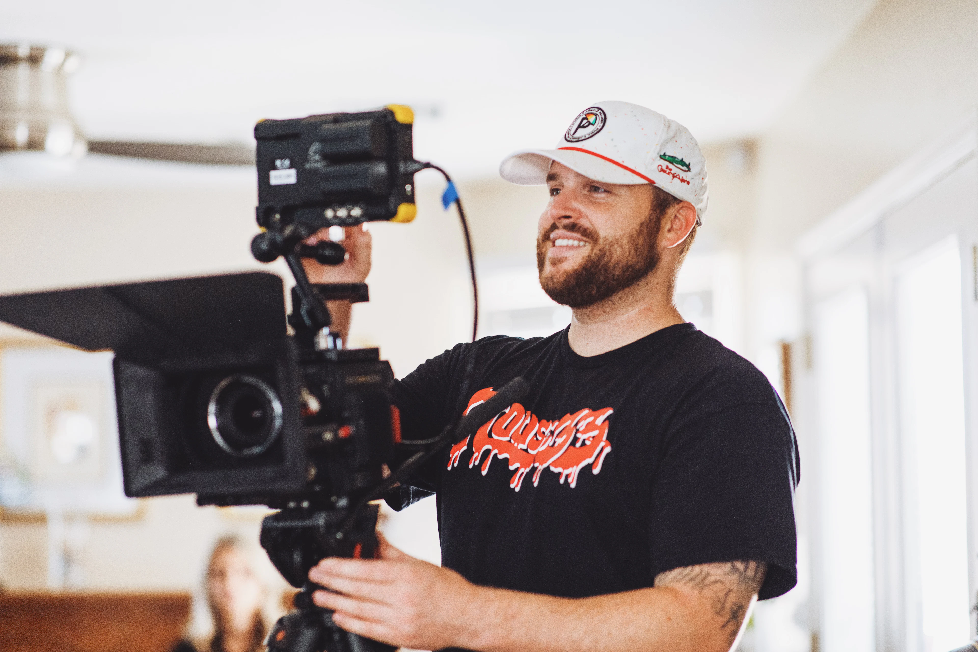 A smiling camera operator in a cap standing behind a camera rig