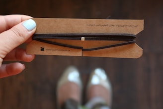 A hand with light blue nail polish is holding a brown cardboard card with a string wrapped around it. The card has printed text in a language other than English. In the background, a pair of blurred shoes is visible, suggesting the photo was likely taken from above.