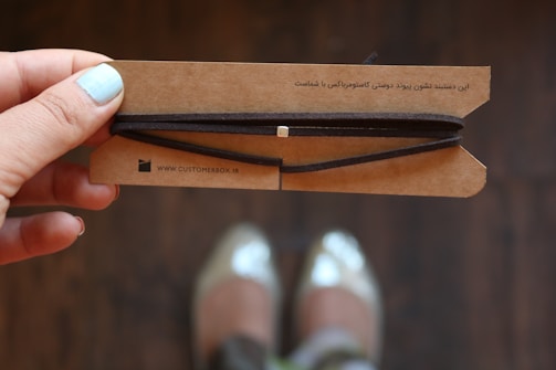 A hand with light blue nail polish is holding a brown cardboard card with a string wrapped around it. The card has printed text in a language other than English. In the background, a pair of blurred shoes is visible, suggesting the photo was likely taken from above.