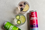 Two cans of Creative Waters Co. cannabis-infused sparkling water are positioned beside two glasses filled with different beverages. One glass contains slices of cucumber and lime in clear liquid, while the other has blackberries and a slice of lemon floating in it. The cans are labeled with different flavors: Lemon-Lime and Berry-Lemon.