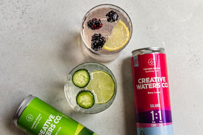 Two cans of Creative Waters Co. cannabis-infused sparkling water are positioned beside two glasses filled with different beverages. One glass contains slices of cucumber and lime in clear liquid, while the other has blackberries and a slice of lemon floating in it. The cans are labeled with different flavors: Lemon-Lime and Berry-Lemon.
