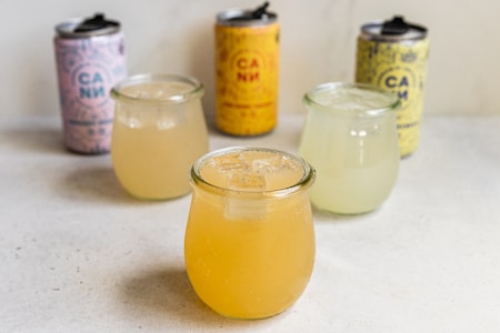 Four glass jars are filled with different colored beverages on a light surface. In the foreground, a glass with a peach-colored drink and ice cubes. In the background, three cans with colorful designs are placed, one pink, one yellow, and another with a blend of colors.