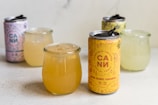 On a light-colored surface, there are several cans and glasses filled with beverages. In the foreground is a can labeled 'Cannabis-infused social tonic' alongside a drink in a clear glass jar. Additional cans and glass jars are placed in the background, including a pink can with the same branding.