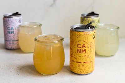 On a light-colored surface, there are several cans and glasses filled with beverages. In the foreground is a can labeled 'Cannabis-infused social tonic' alongside a drink in a clear glass jar. Additional cans and glass jars are placed in the background, including a pink can with the same branding.