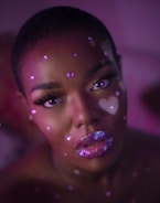 A close-up portrait of an individual with a creative makeup design. The face is adorned with sparkling pink and purple gems and the lips are covered with multicolored glitter. The lighting casts a soft, ethereal glow in shades of pink and purple, adding to the artistic and surreal look.