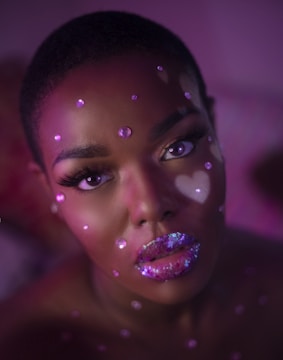 A close-up portrait of an individual with a creative makeup design. The face is adorned with sparkling pink and purple gems and the lips are covered with multicolored glitter. The lighting casts a soft, ethereal glow in shades of pink and purple, adding to the artistic and surreal look.