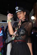 A person wearing a fashionable black outfit with sheer fabric and floral detailing. They have a floral scarf around their neck and a bejeweled wide-brimmed hat. The person is posing confidently indoors with various colorful garments hanging in the background.