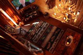 A cozy living room with warm wooden tones is viewed from above. A patterned rug covers the floor, surrounded by rustic wooden furniture including a rocking chair and a wooden table with a candle centerpiece. On the left, a flat-screen TV displays a person's face. An antler chandelier provides warm lighting, and a woven basket sits near a small bookshelf.