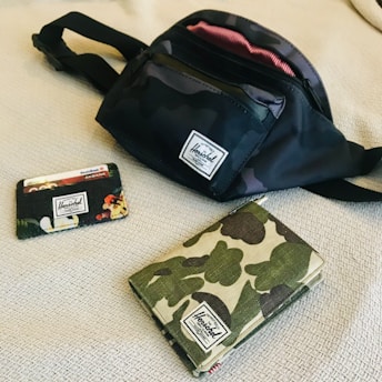 A black and camo-style Herschel fanny pack is placed on a textured, light-colored surface. Nearby, a camouflage-patterned wallet and a cardholder with a floral design are arranged. Each item prominently features the Herschel brand label.