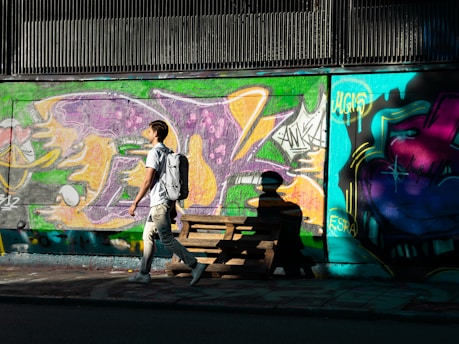 A person with a backpack walks along a street lined with colorful graffiti art on the wall. The artwork features vibrant splashes of purple, green, orange, and blue, with various abstract symbols and tags. The scene is bathed in sunlight, casting strong shadows on the ground and wall.