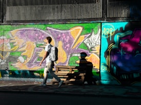 A person with a backpack walks along a street lined with colorful graffiti art on the wall. The artwork features vibrant splashes of purple, green, orange, and blue, with various abstract symbols and tags. The scene is bathed in sunlight, casting strong shadows on the ground and wall.