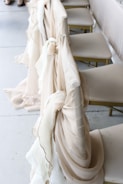 Several chairs are adorned with sheer, flowing fabric in soft beige and cream hues, elegantly draped over the backs. The arrangement suggests a setting for a formal event, possibly a wedding or banquet. The fabric adds a touch of sophistication and luxury to the seating arrangement.