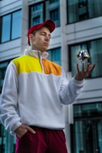 A person wearing a white, yellow, and orange jacket and a red cap is skillfully tossing a Rubik's cube in the air. The background features a modern building with large windows, adding an urban setting to the scene.