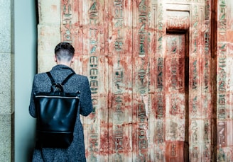 A person with a short haircut stands facing an ancient wall covered in Egyptian hieroglyphs. The individual wears a dark coat and carries a large black leather backpack. The wall's faded sandstone surface is adorned with intricate carvings, showcasing a mix of red and beige colors.