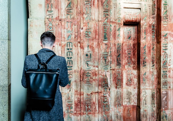 A person with a short haircut stands facing an ancient wall covered in Egyptian hieroglyphs. The individual wears a dark coat and carries a large black leather backpack. The wall's faded sandstone surface is adorned with intricate carvings, showcasing a mix of red and beige colors.