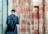 A person with a short haircut stands facing an ancient wall covered in Egyptian hieroglyphs. The individual wears a dark coat and carries a large black leather backpack. The wall's faded sandstone surface is adorned with intricate carvings, showcasing a mix of red and beige colors.