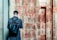 A person with a short haircut stands facing an ancient wall covered in Egyptian hieroglyphs. The individual wears a dark coat and carries a large black leather backpack. The wall's faded sandstone surface is adorned with intricate carvings, showcasing a mix of red and beige colors.