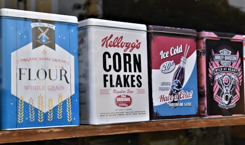 A row of colorful vintage-style metal tins is displayed. Each tin features distinct branding and designs: one blue with 'Flour' labeled on it, another white with bold black 'Kellogg's Corn Flakes' text, a red and blue one advertising cola, and the last one has a Harley-Davidson theme with pink and black hues.