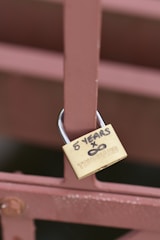 A brass padlock is attached to a brown metal fence. The padlock has the words '5 YEARS x ∞' written in black marker on it, along with the brand name 'TRUGUARD'.