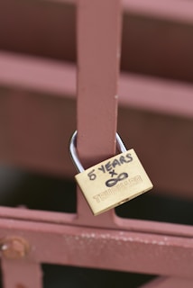 A brass padlock is attached to a brown metal fence. The padlock has the words '5 YEARS x ∞' written in black marker on it, along with the brand name 'TRUGUARD'.