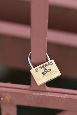 A brass padlock is attached to a brown metal fence. The padlock has the words '5 YEARS x ∞' written in black marker on it, along with the brand name 'TRUGUARD'.
