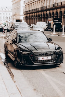 A sleek black car is parked on a city street alongside other vehicles. The car features modern design elements and a distinguishable logo on the front grille. The urban backdrop includes historic architecture with tall columns and arches, contributing to a refined atmosphere.