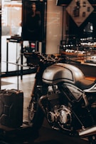 A sleek, silver Norton Commando 961 motorcycle is prominently displayed inside a showroom or a shop. The motorcycle has black detailing and chrome components, emphasized by the lighting. In the background, a black and white photograph, possibly an advertisement, is partially visible along with some shelves containing various items. A metal gas can is placed on the floor near the motorcycle.