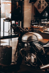 A sleek, silver Norton Commando 961 motorcycle is prominently displayed inside a showroom or a shop. The motorcycle has black detailing and chrome components, emphasized by the lighting. In the background, a black and white photograph, possibly an advertisement, is partially visible along with some shelves containing various items. A metal gas can is placed on the floor near the motorcycle.