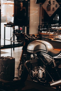 A sleek, silver Norton Commando 961 motorcycle is prominently displayed inside a showroom or a shop. The motorcycle has black detailing and chrome components, emphasized by the lighting. In the background, a black and white photograph, possibly an advertisement, is partially visible along with some shelves containing various items. A metal gas can is placed on the floor near the motorcycle.