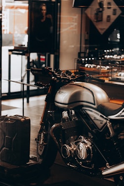A sleek, silver Norton Commando 961 motorcycle is prominently displayed inside a showroom or a shop. The motorcycle has black detailing and chrome components, emphasized by the lighting. In the background, a black and white photograph, possibly an advertisement, is partially visible along with some shelves containing various items. A metal gas can is placed on the floor near the motorcycle.