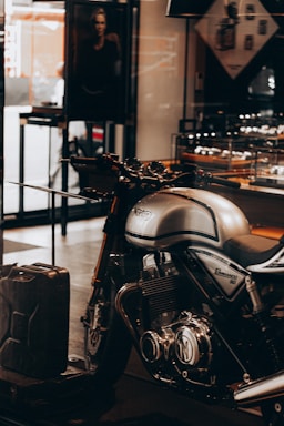 A sleek, silver Norton Commando 961 motorcycle is prominently displayed inside a showroom or a shop. The motorcycle has black detailing and chrome components, emphasized by the lighting. In the background, a black and white photograph, possibly an advertisement, is partially visible along with some shelves containing various items. A metal gas can is placed on the floor near the motorcycle.