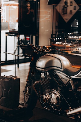 A sleek, silver Norton Commando 961 motorcycle is prominently displayed inside a showroom or a shop. The motorcycle has black detailing and chrome components, emphasized by the lighting. In the background, a black and white photograph, possibly an advertisement, is partially visible along with some shelves containing various items. A metal gas can is placed on the floor near the motorcycle.