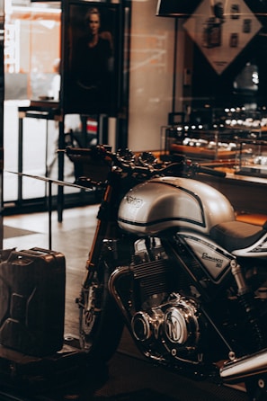 A sleek, silver Norton Commando 961 motorcycle is prominently displayed inside a showroom or a shop. The motorcycle has black detailing and chrome components, emphasized by the lighting. In the background, a black and white photograph, possibly an advertisement, is partially visible along with some shelves containing various items. A metal gas can is placed on the floor near the motorcycle.