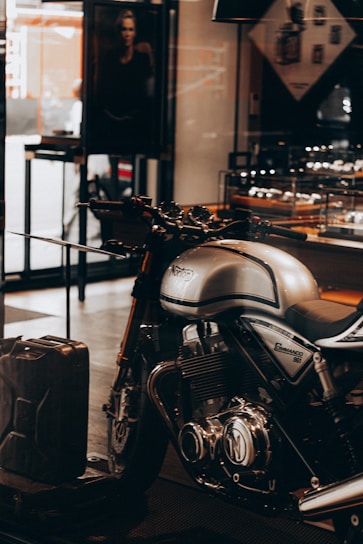 A sleek, silver Norton Commando 961 motorcycle is prominently displayed inside a showroom or a shop. The motorcycle has black detailing and chrome components, emphasized by the lighting. In the background, a black and white photograph, possibly an advertisement, is partially visible along with some shelves containing various items. A metal gas can is placed on the floor near the motorcycle.