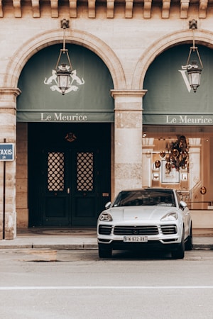 A luxury car is parked in front of the elegant facade of the Le Meurice building, characterized by two large arched windows and ornate lanterns. The building has a classic architectural style with stone columns.