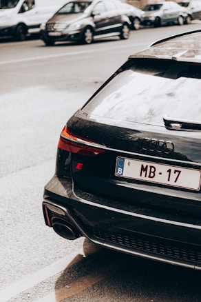 A sleek black Audi car is parked on the side of a busy street. The rear of the car is visible, showing a European license plate with the code 'B' and characters 'MB-17'. Other cars are driving past in the background, indicating a city environment.