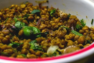 A close-up of a savory dish consisting of cooked chickpeas mixed with various vegetables and garnished with fresh herbs. The chickpeas are golden brown, and there are visible slices of onions and pieces of leafy cilantro. A whole green chili pepper is prominently displayed on top, adding vibrant color to the dish.
