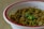 A white bowl with a red rim containing a dish made of cooked chickpeas. The chickpeas are seasoned and garnished with fresh cilantro leaves and spices. The dish appears to have green chili and onions mixed in for flavor.